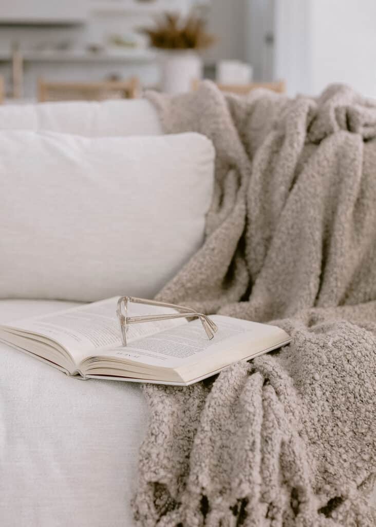Reading glasses resting on an open book beside a soft throw.